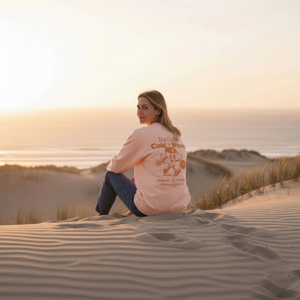 <img src="Hot-Coffee-Cold-Waves-Sweatshirt-fraiche-peche-back-woman-sit-beach_1024px.webp" alt="Frau sitzt in den Dünen am Strand, trägt das Hot Coffee Cold Waves Sweatshirt in Fraîche Pêche: Rückansicht mit Blick über die Schulter in die Kamera, großer orangefarbener Backprint, natürliche Küstenatmosphäre, gefertigt aus 100% GOTS-zertifizierter Bio-Baumwolle">