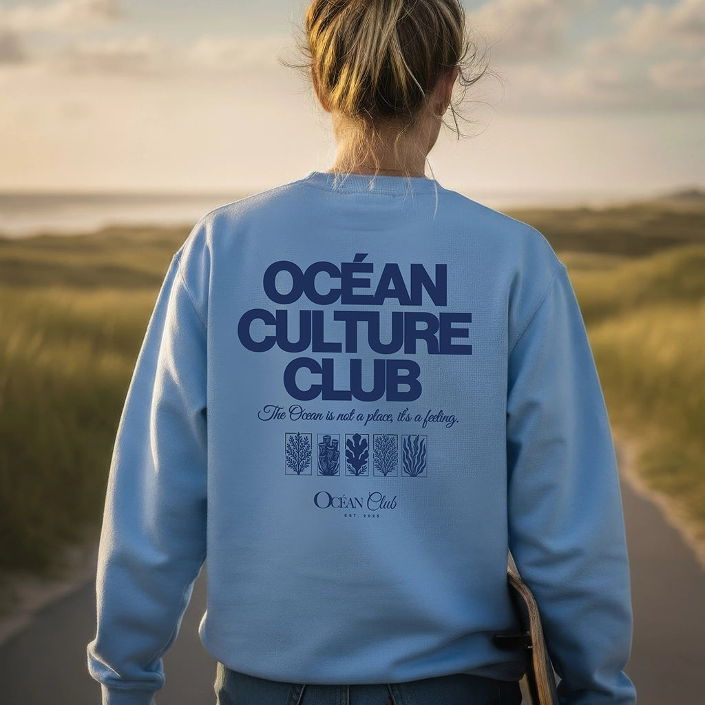 <img src="Ocean-Culture-Club-Sweatshirt-blue-soul-back-woman-walking-Longboard-1024px.webp" alt="Frau in den Dünen mit Longboard unter dem Arm, trägt das Ocean Culture Club Sweatshirt in Blue Soul (hellblau): Close-Up des großen Backprints mit Schriftzug 'Ocean Culture Club' und gradlinigen Illustrationen von Wasserpflanzen, Algen und Korallen darunter, Blick in die Ferne, entspannte Küstenatmosphäre, gefertigt aus 100% GOTS-zertifizierter Bio-Baumwolle">