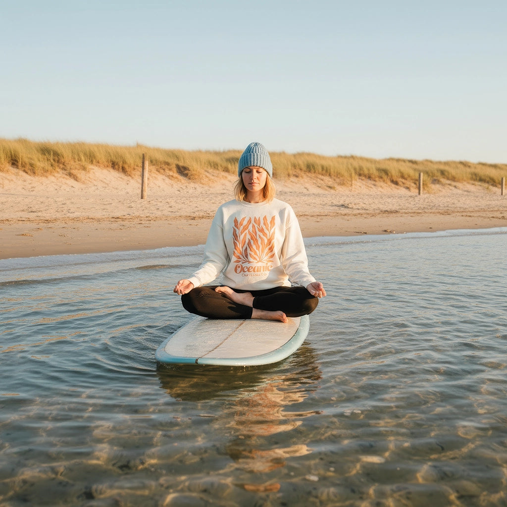 <img src="Oceanic-Club-Sweatshirt-offwhite-front-woman-sup-yoga-lotus-pose_1024px.webp" alt="Frau im Lotussitz auf einem Stand-Up-Paddleboard, trägt das Oceanic Club Sweatshirt in Off-White mit großem Algen-Print in ausgeblichenem Orange auf der Front, meditative Atmosphäre auf ruhigem Wasser, gefertigt aus 100% GOTS-zertifizierter Bio-Baumwolle">