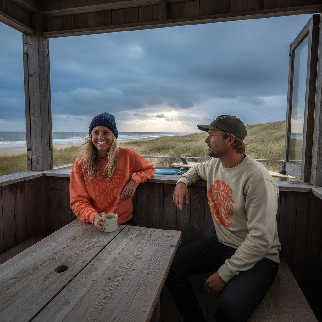 <img src="Tide-and-Time-Sweatshirt-fiesta-stone-front-woman-man-Inside-Beachclub-Cafe-together_1024px.webp" alt="Frau und Mann sitzen entspannt im Strandcafé: Sie hält ein Heißgetränk, beide tragen das Tide and Time Sweatshirt – sie in Fiesta (rot-orange), er in Stone – mit kreisförmigem Frontprint (stilisierte Welle und Schriftzug 'Nordic Soulmates'), gemütliche Café-Atmosphäre, gefertigt aus 100% GOTS-zertifizierter Bio-Baumwolle">