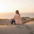 <img src="Hot-Coffee-Cold-Waves-Sweatshirt-fraiche-peche-back-woman-sit-beach_1024px.webp" alt="Frau sitzt in den Dünen am Strand, trägt das Hot Coffee Cold Waves Sweatshirt in Fraîche Pêche: Rückansicht mit Blick über die Schulter in die Kamera, großer orangefarbener Backprint, natürliche Küstenatmosphäre, gefertigt aus 100% GOTS-zertifizierter Bio-Baumwolle">