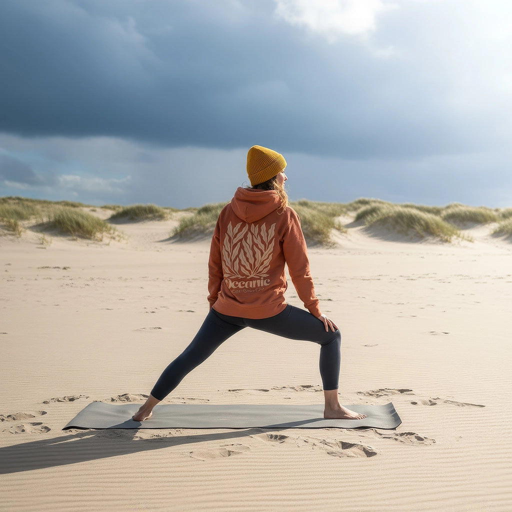 <img src="Oceanic-Club-Hoodie-heritage-brown-back-woman-Yoga-sport-high-lunge_1024px.webp" alt="Frau in High Lunge am Strand, trägt den unisex Oceanic Club Hoodie in Heritage Brown: großer Algen-Backprint, dynamische Yoga-Pose mit Meerblick, gefertigt aus 100% GOTS-zertifizierter Bio-Baumwolle">