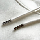 <img src="weisser-hoodie-struktur-biobaumwolle-doppelnaht-kordeln-detail.jpg" alt="Detailaufnahme eines weißen Hoodies: feine Struktur der gekämmten, ringgesponnenen Bio-Baumwolle, präzise Doppelnaht-Steppstiche an Ärmelbündchen und Saum, hochwertige Kordeln mit Metallenden, gefertigt aus 100% GOTS-zertifizierter Bio-Baumwolle">