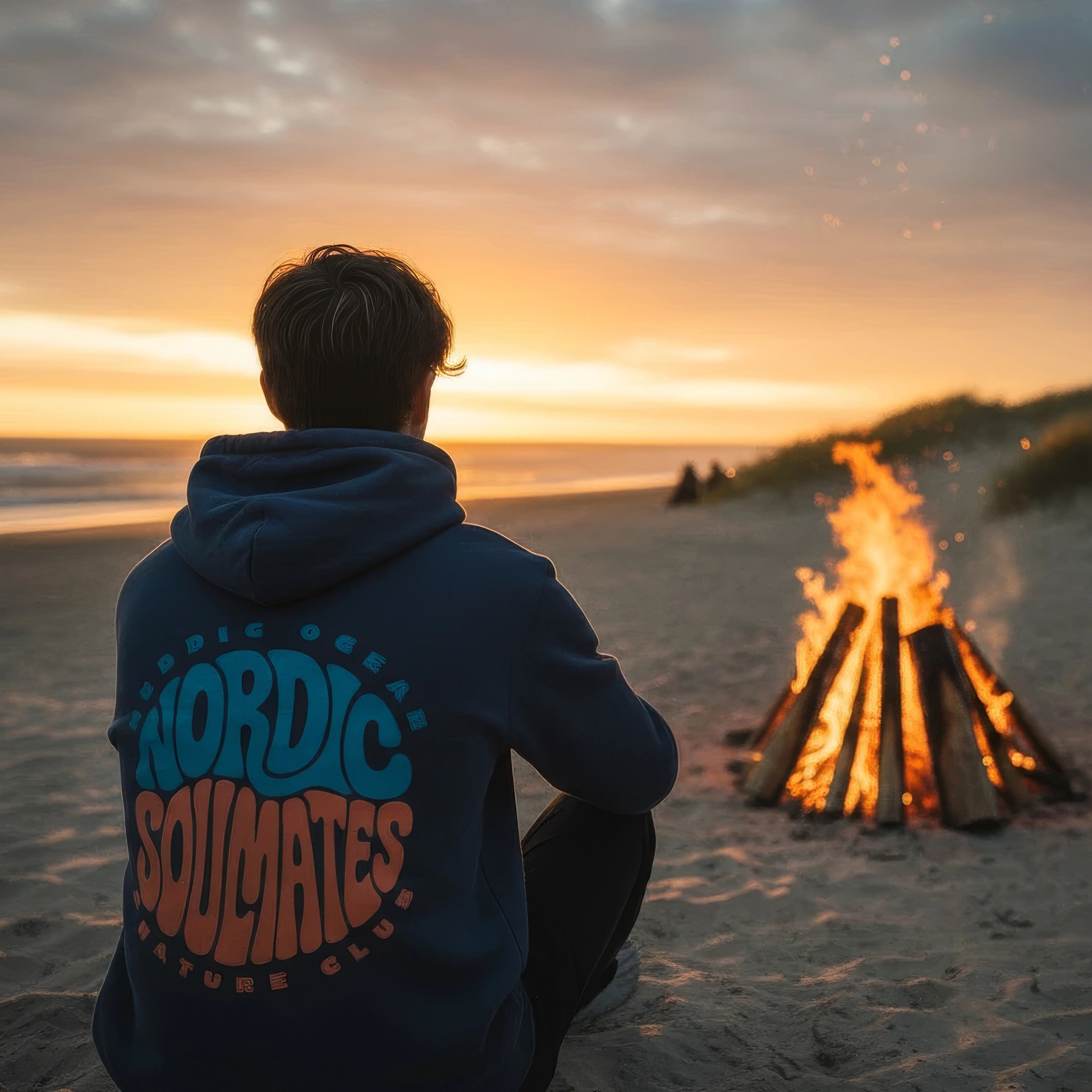 <img src="unity-circle-hoodie-lagerfeuer.jpg" alt="Mann trägt den Unisex Nordic Soulmates Unity Circle Hoodie in India Ink Grey am Lagerfeuer am Strand: Rückansicht mit großem Backprint, umhüllt von warmem Feuerlicht und rauer Küstenatmosphäre, gefertigt aus 100% GOTS-zertifizierter Bio-Baumwolle">